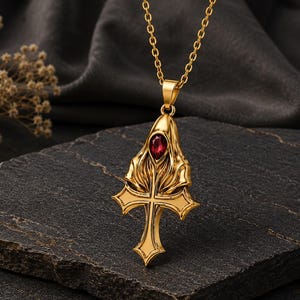 Könnte beinhalten: Eine goldfarbene Anhängerkette mit einem Kreuzdesign, das oben mit kunstvollen, fließenden Metallverzierungen versehen ist und in der Mitte einen einzelnen, tropfenförmigen roten Edelstein trägt. Der Anhänger hängt an einer passenden goldfarbenen Gliederkette.