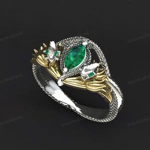 以下が含まれることがあります： マーキス型のエメラルド宝石をあしらった、シルバーとゴールドのデザインが特徴的な詳細なリング。リングは、小さな宝石やテクスチャ加工された金属細工など、複雑なディテールを持つヘビのようなモチーフです。
