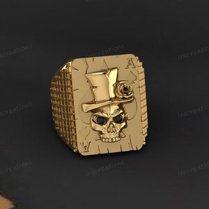 May include: A gold-toned ring featuring a rectangular face shaped like an ace of spades playing card. The design includes a raised skull wearing a top hat with a small rose detail, and the skull has two small black stones set as eyes.