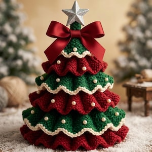 Puede incluir: Un adorno de árbol de Navidad tejido en niveles, con capas alternas de tela verde oscuro y rojo intenso con bordes ondulados. El árbol está decorado con pequeñas cuentas de perlas blancas, rematado con una estrella plateada y un gran lazo de satén rojo.