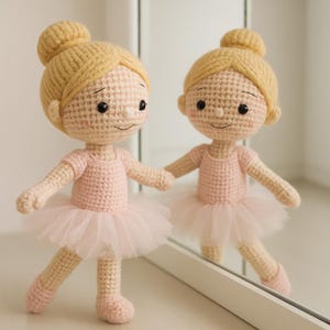May include: Two crochet ballerina dolls with blonde buns, wearing pink leotards and tulle skirts. The dolls have black eyes and are standing in front of a mirror. The dolls are handmade and have a whimsical appearance.