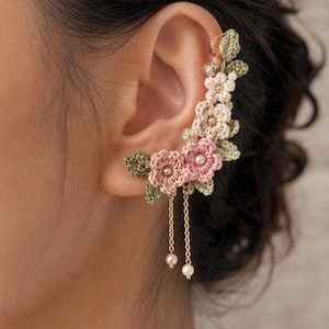Può includere: Un orecchino a crochet con un design floreale nei toni del rosa, crema e verde. Il polsino include delicate catene dorate con perle finte pendenti. I fiori sono finemente dettagliati, creando un accessorio unico ed elegante.
