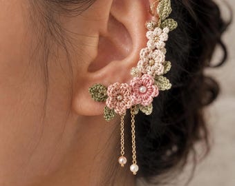 May include: A crochet ear cuff featuring a floral design in shades of pink, cream, and green. The cuff includes delicate gold chains with dangling faux pearls. The flowers are intricately detailed, creating a unique and elegant accessory.
