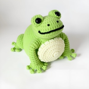 May include: A handmade crochet frog in shades of green and cream. The frog has large black eyes, a black smile, and detailed feet. The belly is a cream color, and the frog is sitting upright.