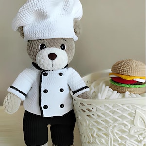 May include: A handmade teddy bear dressed as a chef, wearing a white chef's hat and jacket with black trim and buttons, and black pants. A crocheted hamburger sits in a white basket.