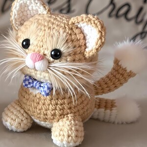 Może przedstawiać: Ręcznie robiona szydełkowa lalka kota w odcieniach beżu i kremu, z niebiesko-białą kraciastą muszką. Kot ma czarne oczy, różowy nos i długie białe wąsy. Ogon i końcówki uszu są białe.