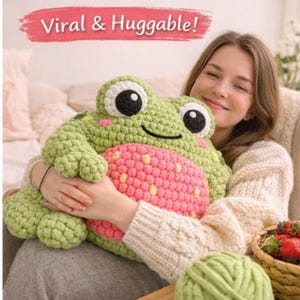 May include: A large, handmade crochet frog pillow in shades of green and pink. The frog has large black and white eyes and a smiling mouth. The text "Viral & Huggable!" is displayed above the pillow in a pink banner.