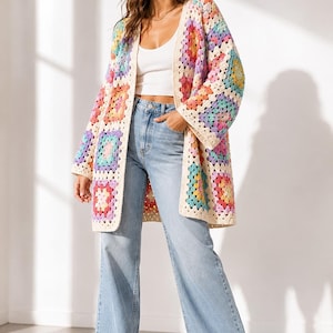 May include: A colorful, open-front crochet cardigan with a granny square design. The long-sleeved sweater features squares in shades of pink, purple, blue, yellow, and green, with a cream-colored border. The cardigan is paired with light blue jeans.