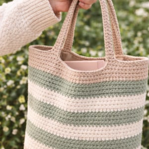 May include: A crocheted tote bag with horizontal stripes in beige, green, and off-white. The bag has two handles and a solid beige bottom. The bag is held up by a person. The background is blurred green foliage.