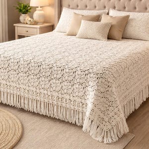 Puede incluir: Un cubrecama color crema con un patrón floral de ganchillo y flecos. La cama está hecha con varias almohadas en tonos neutros. La habitación tiene una paleta de colores neutros con una alfombra redonda tejida.