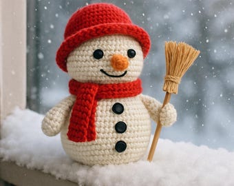 May include: A crocheted snowman with a red hat, scarf, and black button details. It has a carrot nose and a smiling face. The snowman is holding a small broom and is sitting on a snowy surface.