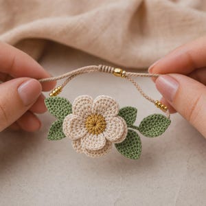 May include: A handmade crochet bracelet featuring a cream-colored flower with a yellow center and green leaves. The bracelet has a tan cord with gold-colored beads. The flower is approximately 2 inches in diameter.