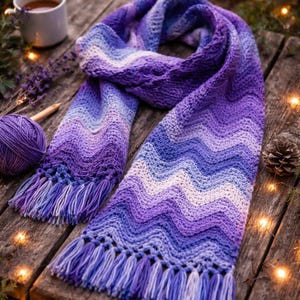 May include: A handmade, chevron-patterned scarf in shades of purple, lavender, and white, with fringe at the ends. The scarf is displayed on a rustic wooden surface, with a ball of yarn and a cup of tea nearby. The scarf is approximately 72 inches long.