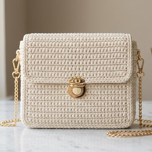 May include: Cream-colored crochet crossbody bag with a rectangular shape and a flap closure. The bag features a gold-colored clasp and chain strap. A small vase with dried leaves is in the background.