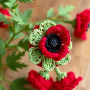 May include: Crocheted red poppy flowers with green accents and black centers. The handmade flowers are attached to green stems with green leaves. The flowers are arranged on a wooden surface.