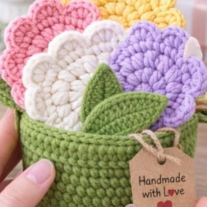 Op de afbeelding: Een handgemaakt gehaakt bloemstuk in een groene pot. De bloemen zijn in tinten roze, wit, geel en paars, met groene bladeren. Een klein label leest "Handmade with Love" met een rood hart.