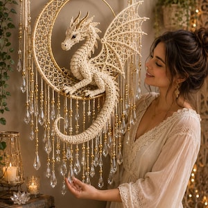 Op de afbeelding: Een crèmekleurige macramé wandhanger met een centrale drakenfiguur op een gouden halve maan ring. Het stuk hangt aan een houten tak en is versierd met hangende gouden kralen en heldere, kristalachtige druppelvormige hangers.