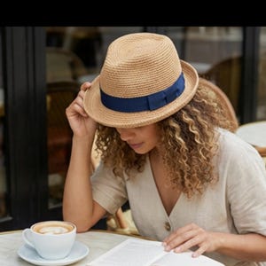 May include: A woven straw fedora hat with a dark blue band. The hat is worn by a person sitting at a table with a cup of coffee and a book. The hat's brim is slightly curved, and the band adds a pop of color.