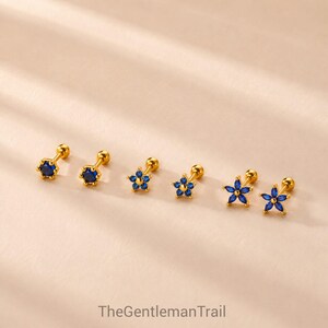 Può includere: Tre paia di orecchini a lobo color oro con pietre blu. Il primo paio presenta una pietra blu rotonda, il secondo un design a fiore a sei petali e il terzo un fiore a sei petali allungati. Il testo riporta TheGentlemanTrail.