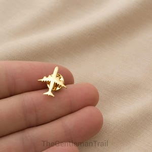 Könnte beinhalten: Eine goldfarbene Anstecknadel aus Metall in Form eines Verkehrsflugzeugs mit schlanker Silhouette, bestehend aus einem zentralen Rumpf, zwei Flügeln und einem Leitwerk. Die Nadel hat eine polierte, reflektierende Oberfläche und einen Verschluss.