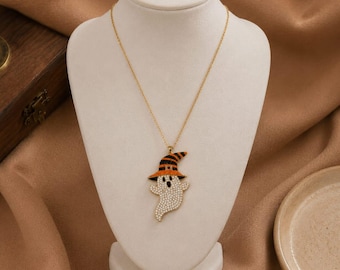 Puede incluir: Un collar de cadena en tono dorado con un colgante de fantasma adornado con cristales transparentes. El fantasma lleva un sombrero de bruja decorado con cristales negros y naranjas. El colgante cuelga de una fina cadena de metal dorado.