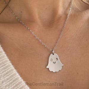 Op de afbeelding: Een zilverkleurige ketting met een platte, spookvormige hanger met uitgesneden ogen en een kleine mond. De hanger hangt aan een fijne zilverkleurige ketting. De tekst "TheGentlemanTrail" is zichtbaar onderaan de afbeelding.