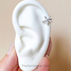 Puede incluir: Un pendiente de oreja plateado con un diseño floral con cristales transparentes y brillantes. El pendiente, con forma de estrella, se lleva en el cartílago de la oreja. El fondo es de color beige neutro.