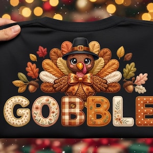Pode incluir: Uma t-shirt preta com um peru de desenho animado usando um chapéu de peregrino e uma gravata borboleta, rodeado por folhas de outono e bolotas. Abaixo do peru, a palavra GOBBLE está impressa em letras grandes com padrões florais e xadrez.