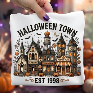 May include: A white fabric square featuring a black and orange illustration of a spooky town with the text HALLOWEEN TOWN and EST 1998. The design includes gothic buildings, a water tower, ghosts, pumpkins, bats, and candy accents.