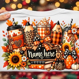 May include: A white t-shirt featuring a colorful autumn-themed graphic with coffee cups, pencils, sunflowers, pumpkins, and books. The design includes a central black sign with the text Name here, surrounded by orange, brown, and black patterns.