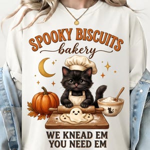 May include: A white crewneck sweatshirt featuring a graphic of a black cat wearing a chef hat and apron, rolling dough shaped like a ghost on a wooden board. The design includes a pumpkin, a crescent moon, and the text SPOOKY BISCUITS bakery WE KNEAD EM YOU NEED EM.