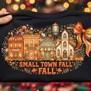May include: A black t-shirt featuring a colorful graphic of a small town street with a bakery, bookshop, general store, and church. The design includes a large orange bow, autumn flowers, pumpkins, and the text "SMALL TOWN FALL".