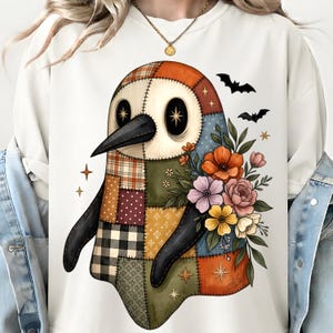 May include: A cream-coloured sweatshirt featuring a graphic of a patchwork-style penguin ghost. The penguin is composed of various plaid, polka dot, and solid fabric patterns in orange, brown, and green, accented with colourful floral bouquets and bats.
