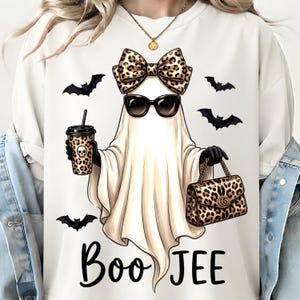 May include: A cream-colored crewneck sweatshirt featuring a graphic of a ghost wearing black sunglasses and a large leopard print bow. The ghost holds a leopard print cup with a skull icon and a leopard print handbag, surrounded by black bats.
