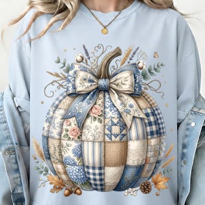 May include: A light blue sweatshirt featuring a large central graphic of a pumpkin composed of various blue and beige patchwork patterns, including floral, gingham, and lace designs, topped with a large blue and white patterned fabric bow.
