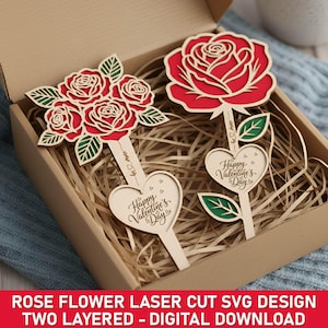 Op de afbeelding: Twee-laags lasergesneden houten rozenbloemdecoraties. Eén heeft een enkele rode roos, de andere een boeket rozen. Beide hebben een hart met "Happy Valentine's Day" en bladeren. De items zitten in een kartonnen doos.