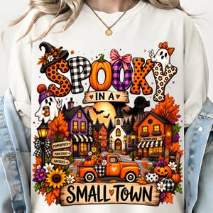May include: A white sweatshirt featuring a colorful graphic with the text SPOOKY IN A SMALL TOWN. The design includes a ghost, an orange pickup truck, pumpkins, sunflowers, and a small town scene with houses, trees, and a full moon background.