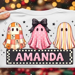 May include: A white t-shirt featuring a graphic of three ghosts. The left ghost has an orange and pink floral patchwork pattern, the centre ghost is pink with white polka dots and a black bow, and the right ghost has orange and white stripes. Below the ghosts is a black rectangular banner with the name AMANDA in pink letters.