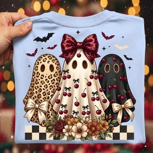 May include: A light blue t-shirt featuring a graphic of three ghosts. The left ghost has a leopard print pattern and a white bow. The center ghost is cream with a large red leopard print bow and cherry motifs. The right ghost is black with cherry motifs and a beige bow. A floral arrangement sits at the base above a black and white checkered border, surrounded by small bats.