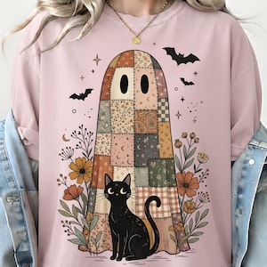 May include: A light pink t-shirt featuring a graphic of a ghost covered in a patchwork quilt pattern, a black cat sitting in front, and surrounding floral elements with small stars and bats. The design uses muted earth tones and black ink.