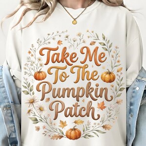 Könnte beinhalten: Ein cremefarbenes Sweatshirt mit Rundhalsausschnitt und einem zentralen Aufdruck mit dem Text "Take Me To The Pumpkin Patch" in brauner Schreibschrift. Der Text ist von einem kreisförmigen Kranz aus orangefarbenen Kürbissen, Herbstblättern und weißen Blumen umgeben.