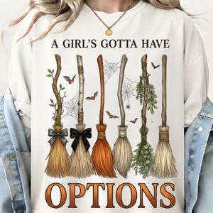 May include: A cream-colored crewneck sweatshirt featuring the text A GIRL'S GOTTA HAVE OPTIONS in black and orange lettering. The graphic displays six distinct witch brooms decorated with bows, greenery, and spiderwebs, accompanied by small flying bats.