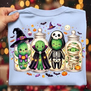 Puede incluir: Una camiseta azul claro con un gráfico de cuatro cactus verdes dentro de frascos de vidrio, disfrazados de bruja, vampiro, esqueleto y momia. El diseño incluye calabazas, murciélagos, dulces y una luna, iluminados por luces de cadena.