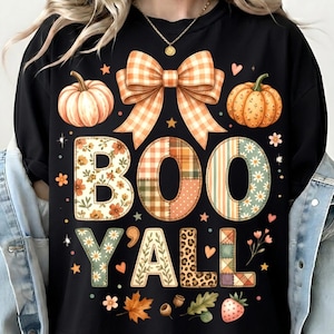 Peut inclure: Un t-shirt noir avec une impression graphique portant le texte "BOO Y'ALL" en grandes lettres à motifs. Le design comprend un grand nœud en vichy orange, deux citrouilles, des petites fleurs, des feuilles, des glands et des étoiles.