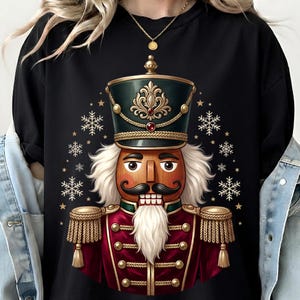 May include: A black t-shirt featuring a graphic of a nutcracker soldier wearing a dark green hat with gold filigree and a red uniform with gold tassels. The design is surrounded by white snowflakes and small gold stars on a black background.