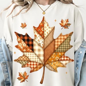 May include: A cream-colored sweatshirt featuring a large central maple leaf graphic composed of various patchwork patterns including orange floral prints, black and orange buffalo plaid, brown herringbone, and tan gingham checks.