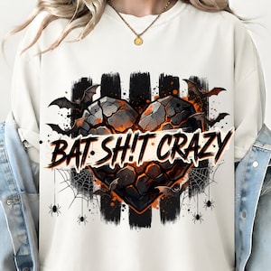 Op de afbeelding: Een crèmekleurig sweatshirt met ronde hals en een afbeelding van een gebarsten, gloeiend oranje en zwart hart. Het ontwerp bevat vliegende vleermuizen, spinnenwebben en spinnen, met de tekst BAT SHIT CRAZY in een vetgedrukt, zwart lettertype.