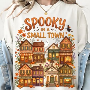 May include: A cream-colored sweatshirt featuring a graphic print with the text SPOOKY IN A SMALL TOWN. The design depicts a collection of small shops including a bakery, bookshop, and apothecary, decorated with pumpkins, flowers, and ghosts.