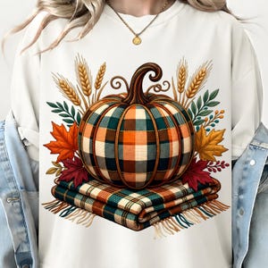 Op de afbeelding: Een wit sweatshirt met ronde hals en een grafische print van een pompoen met een groen, oranje en wit ruitpatroon. De pompoen rust op een gevouwen geruite deken, omringd door illustraties van gouden korenaren en herfstachtige esdoornbladeren.