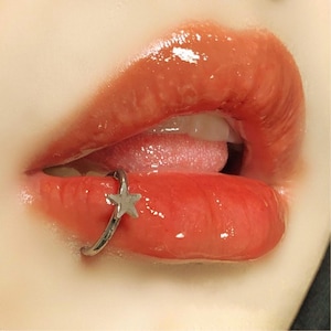 Può includere: Primo piano di labbra rosse lucide con un piercing al labbro argentato con un ciondolo a forma di stella. Le labbra sono socchiuse, rivelando uno scorcio della lingua e dei denti. La pelle intorno alle labbra è liscia e leggermente colorata.